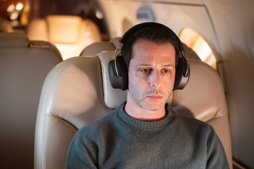 The actor sits in an aeroplane seat with large black headphones and a desolate look on his face.