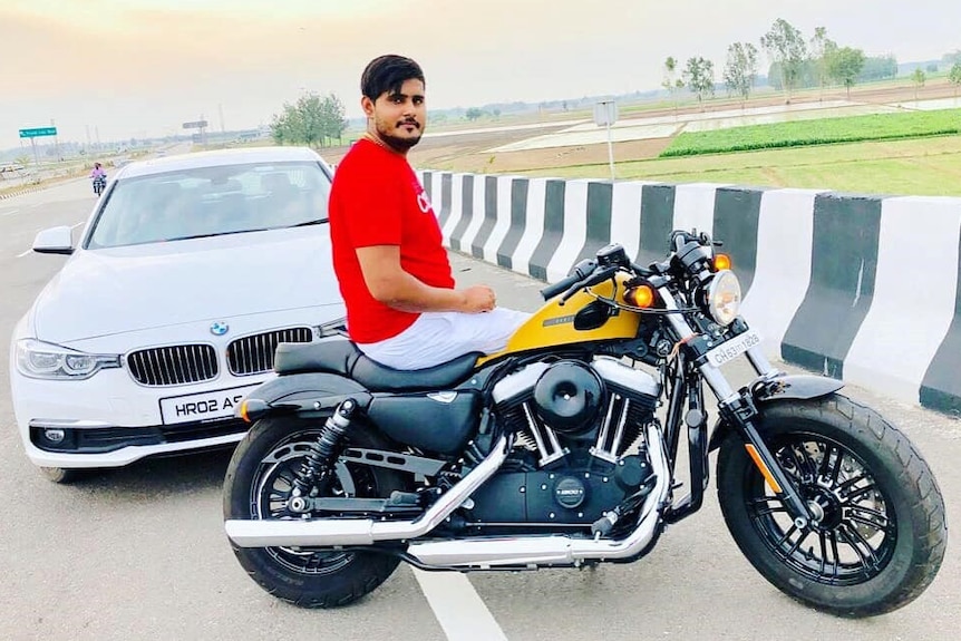 A 22-year-old man from India, known only as Akash can be seen sitting on a motorcycle, in front of a BMW on the side of the road