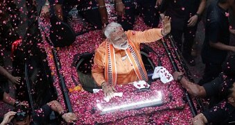 A man in an orange outfit waves from a car that is covered in pink rose petals.