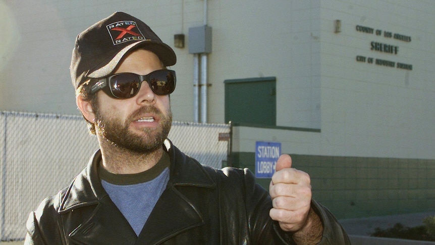 Adam Rich wears a black cap and sunglasses and holds out his fist. 