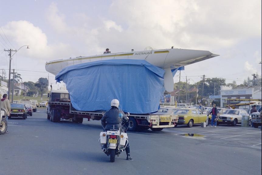 A boat with its keel hidden under a blue tarp being moved on a truck with a police officer on a motorbike