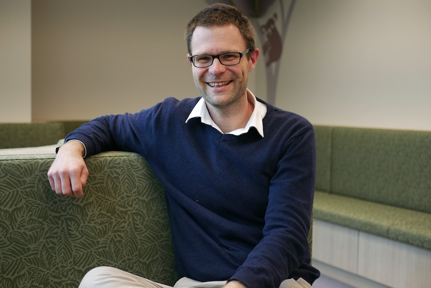 A man wearing glasses and a blue sweater sitting on a green couch.