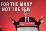 Labour party leader Jeremy Corbyn holds his hands out by his side in a gesture while speaking in front of a red backdrop.