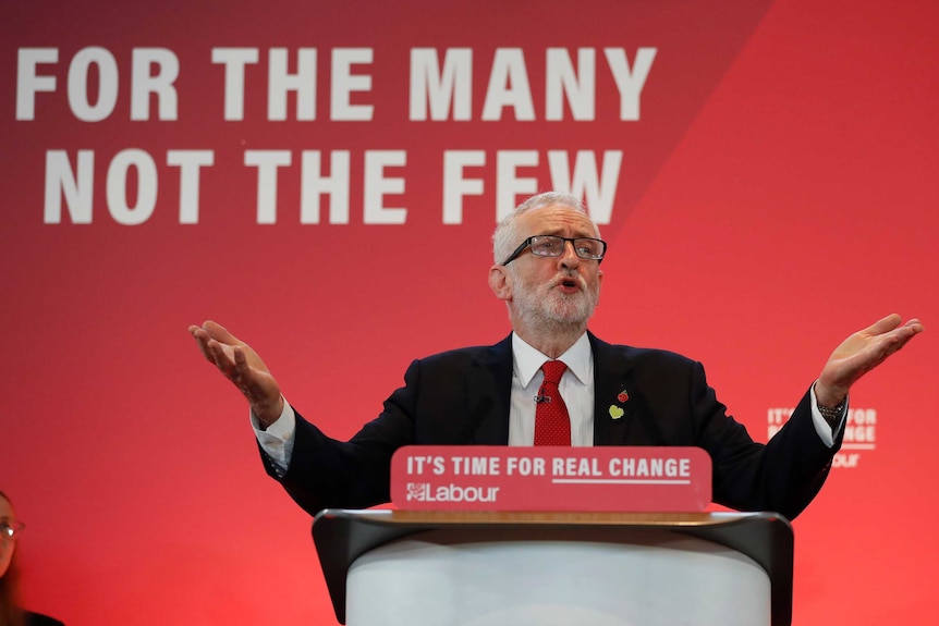 Former Labour leader Jeremy Corbyn holds his hands out by his side in a gesture while speaking in front of a red backdrop.