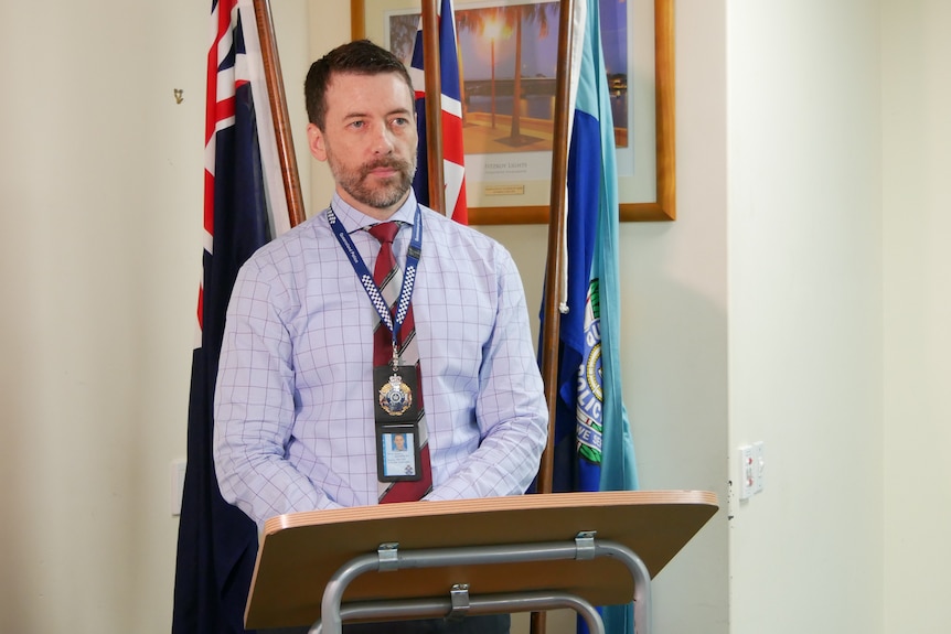 A police officer in plain clothes stands at a lectern, speaking.