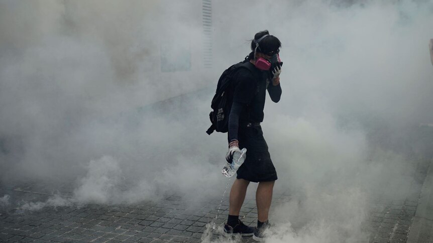 A person in dark clothing with a gas mask on stands in a street surrounded by smoke.