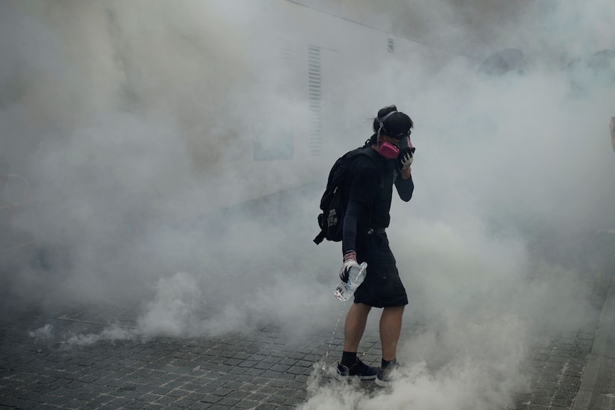 A person in dark clothing with a gas mask on stands in a street surrounded by smoke.