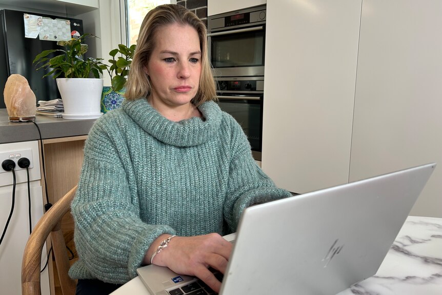 a woman wearing a high neck jumper sitting at a desk typing into her laptop