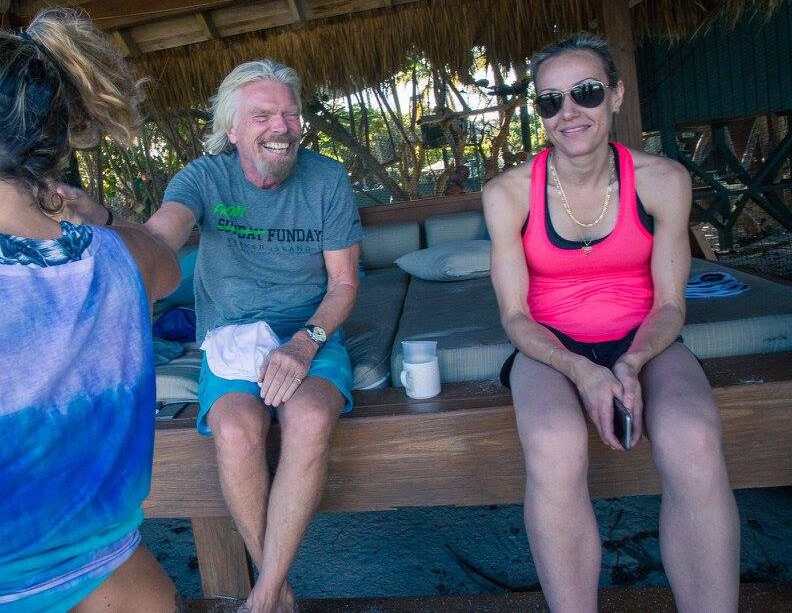 A woman in a pink singlet and sunglasses sits on a thatch-roofed day bed on a beach next to a smiling Richard Branson.