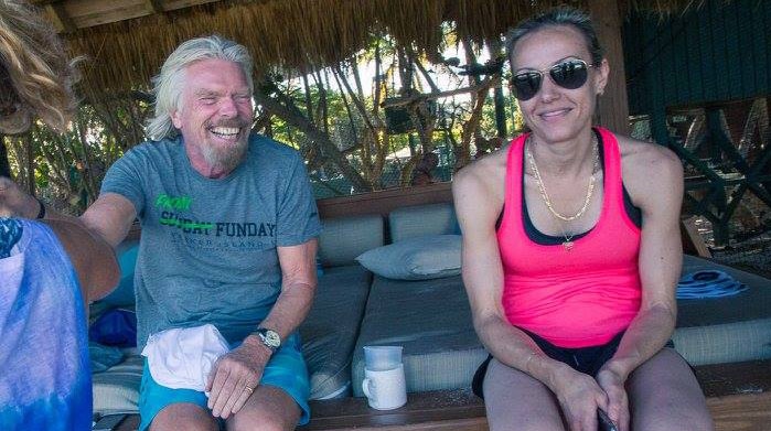 A woman in a pink singlet and sunglasses sits on a thatch-roofed day bed on a beach next to a smiling Richard Branson.