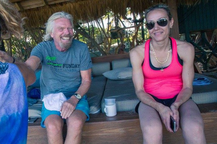 A woman in a pink singlet and sunglasses sits on a thatch-roofed day bed on a beach next to a smiling Richard Branson.