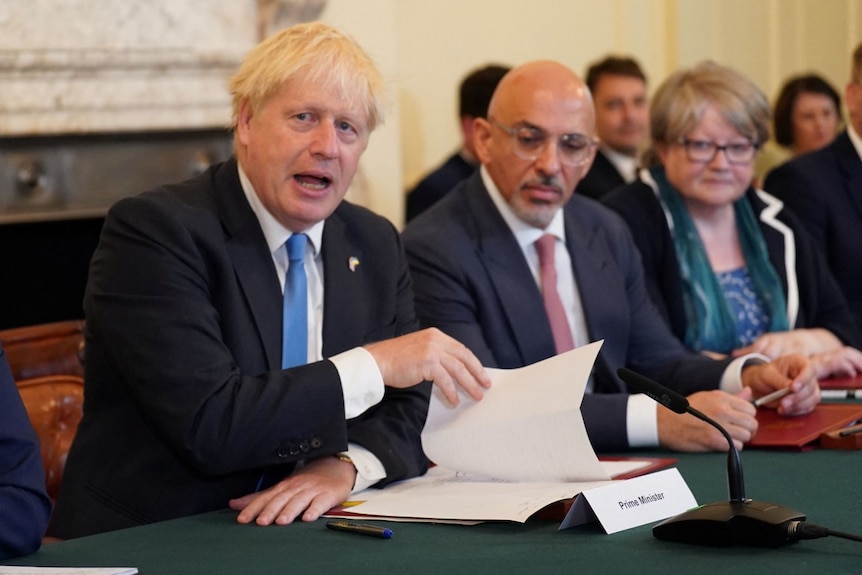 Boris Johnson speaks at a conference table while holding open a folder of papers. Zahawi sits beside him