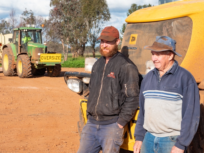 A pair of farmers, one young and one old, lean against a piece of equipment.