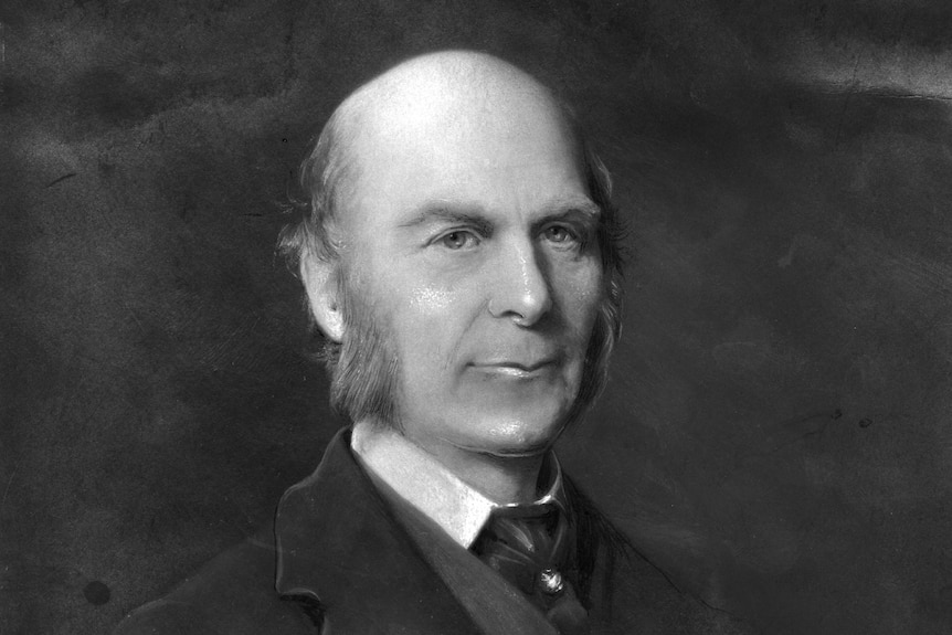 Black and white portrait of a bald man with grey sideburns, wearing a black suit and tie.