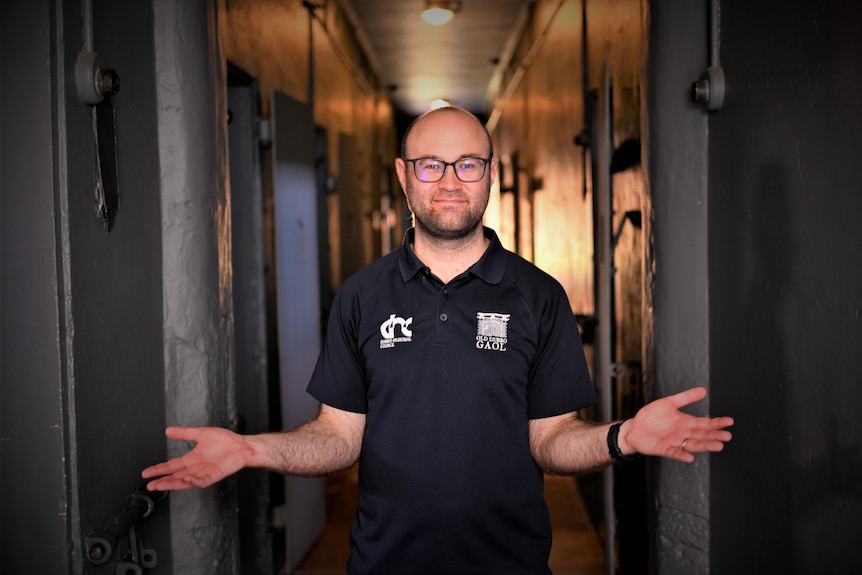 A bespectacled man in a dark polo shirt stands in a corridor in a historic jail.