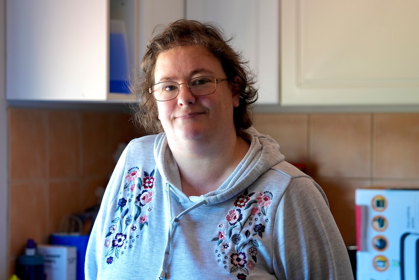 A woman in a grey top stands in a kitchen. 