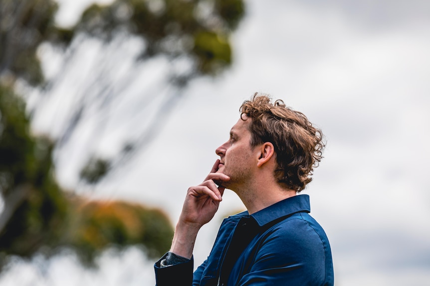 A man holds his hand to his chin and looks up towards a tree