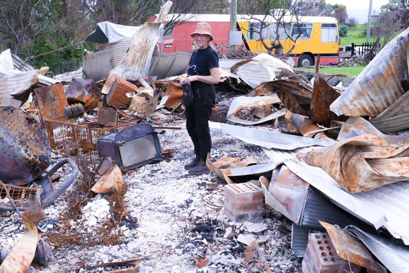 Woman standing amongst rubble with burnt household objects wearing rough work clothes holding gloves