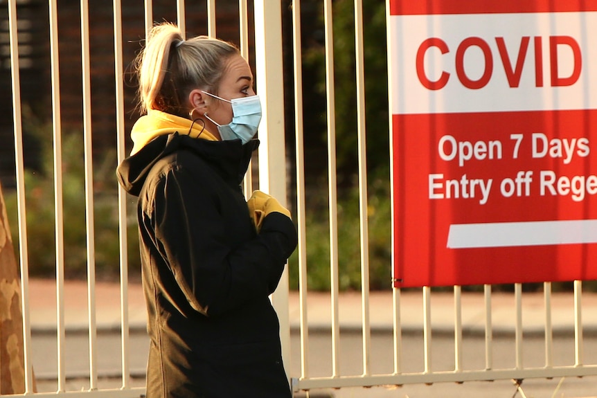 A close-up side-on shot of a woman wearing a face mask in front of a fence with a red COVID clinic sign on it.