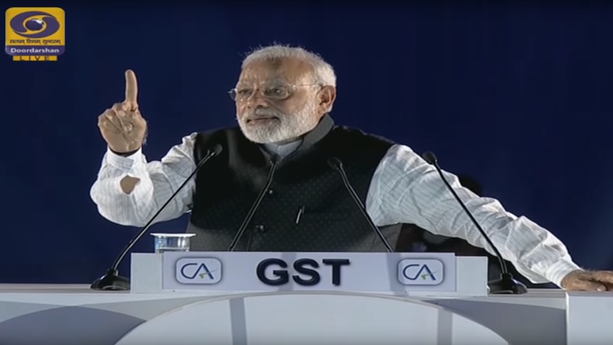 Mr Modi stands at a podium labelled 'GST' with his right hand raised, index finger pointing into the air.