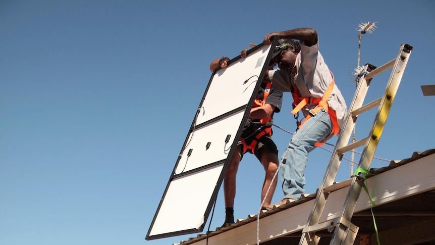 Two men standing on the ground hand a solar panel to two men standing on a rooftop.