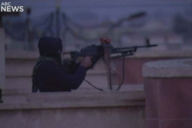 A figure dressed in black stands on a rooftop terrace, aiming a machine gun.