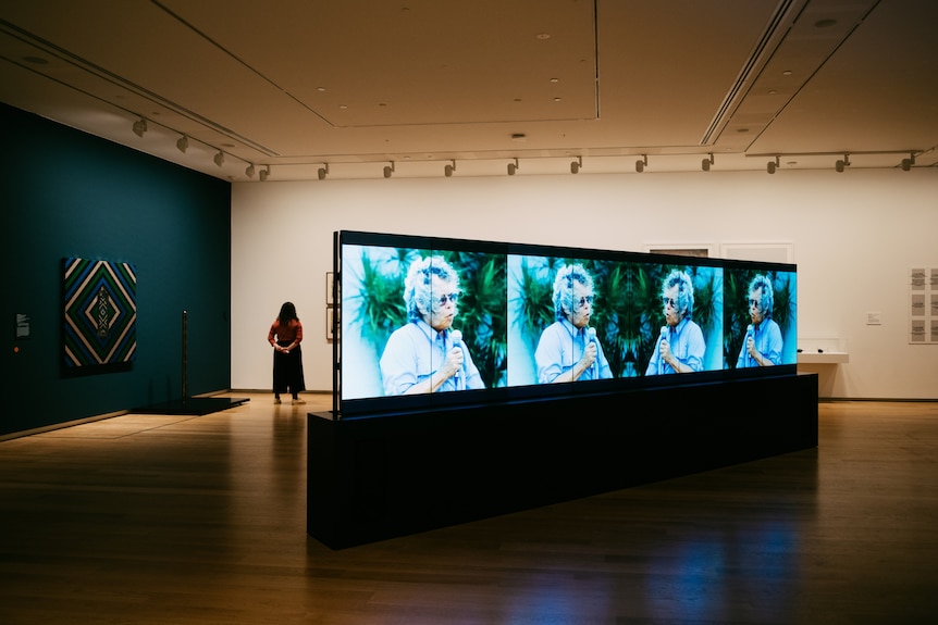 Gallery shot with a four-channel digital video work in foreground, showing an older First Nations woman speaking into a mic.