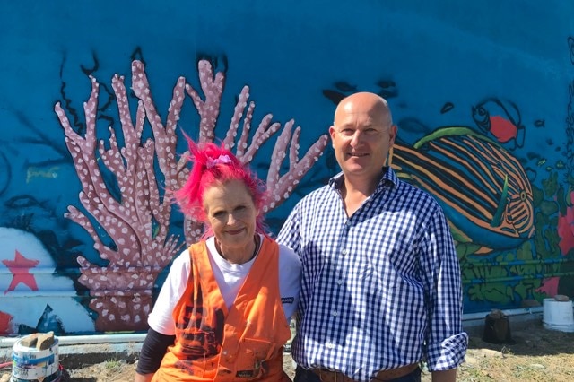 A man and woman stand beside a painted mural of the ocean.