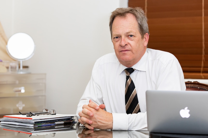 A man in a white shirt with a black and gold tie looks at the camera while sitting at a desk with a glass top.