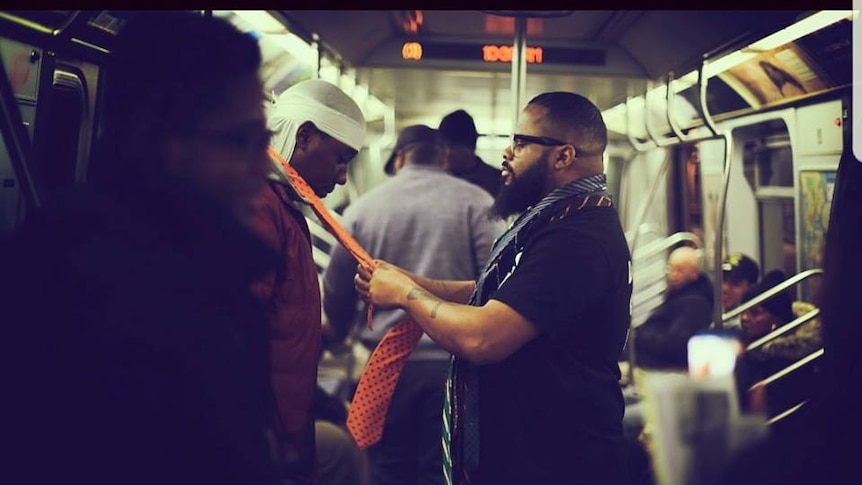 Kevin Livingston of 100 Suits tying a man's tie on a New York subway train.