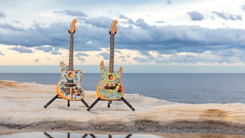 two guitars, with first nations designs on them, standing upright on a sandy beach
