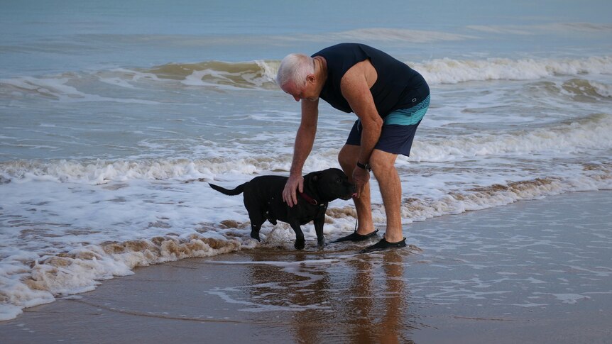 Man leaning over dog at the water's edge.