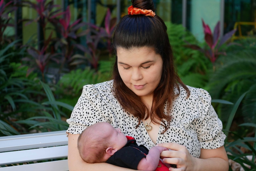 Brunette woman in floral dress looking down at three week old son in her arms at hospital courtyard.