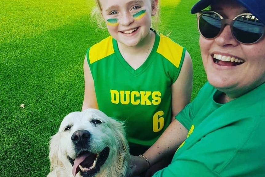 A girl in a sports uniform and a woman in sunglasses and a green shirt hug a white dog