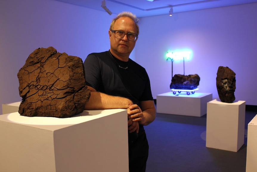 A man in his 50s with glasses and short hair stands in a gallery next a plinth with a piece of coal
