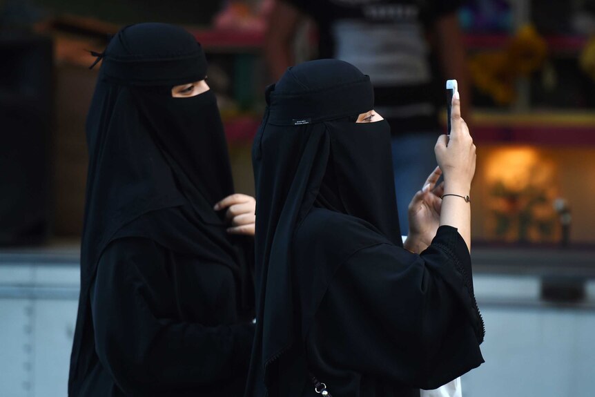 Two women wearing black burkas look at a mobile phone.