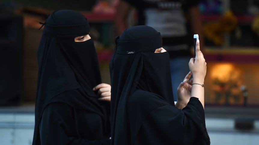 Two women wearing black burkas look at a mobile phone.