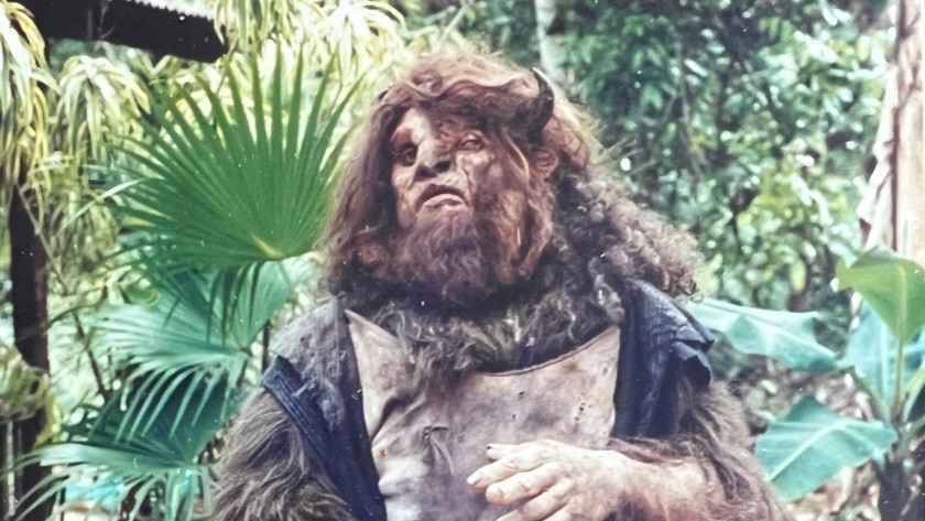 A man in full make-up and prosthetics to look like a human-bison hybrid. with rainforest plants in the background.