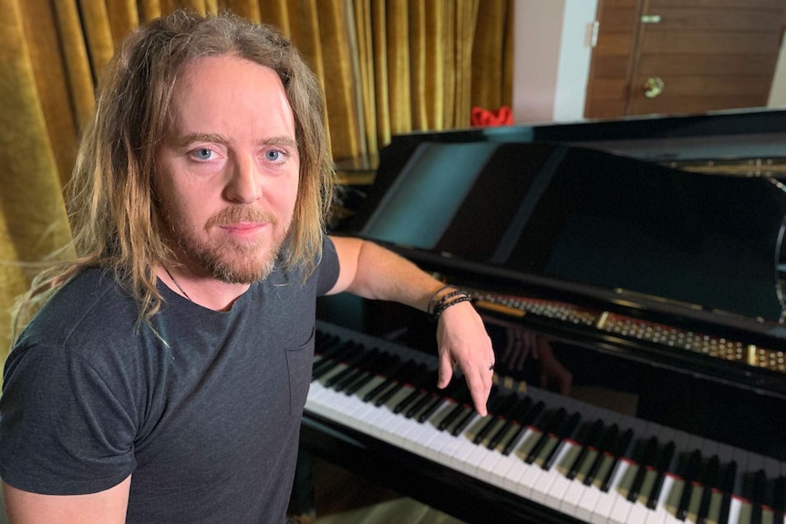 Tim Minchin sits at a grand piano, looking at the camera.