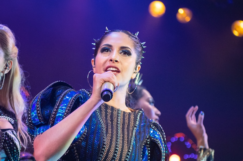 A young woman wearing a silver-spiked headband and blue and black studded dress, holding mic and singing.