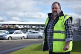 A man wearing a high vis vest over a patterned shirt stands in from of parked cars