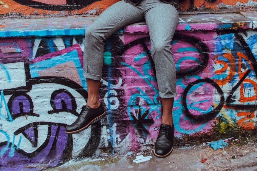 A man wearing shoes without socks sitting against a graffitied wall
