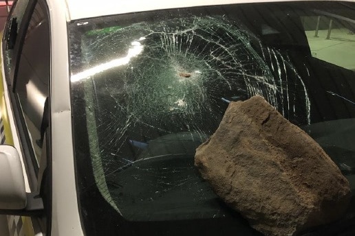A large rock sits on the windscreen of a police car. The windscreen is shattered.