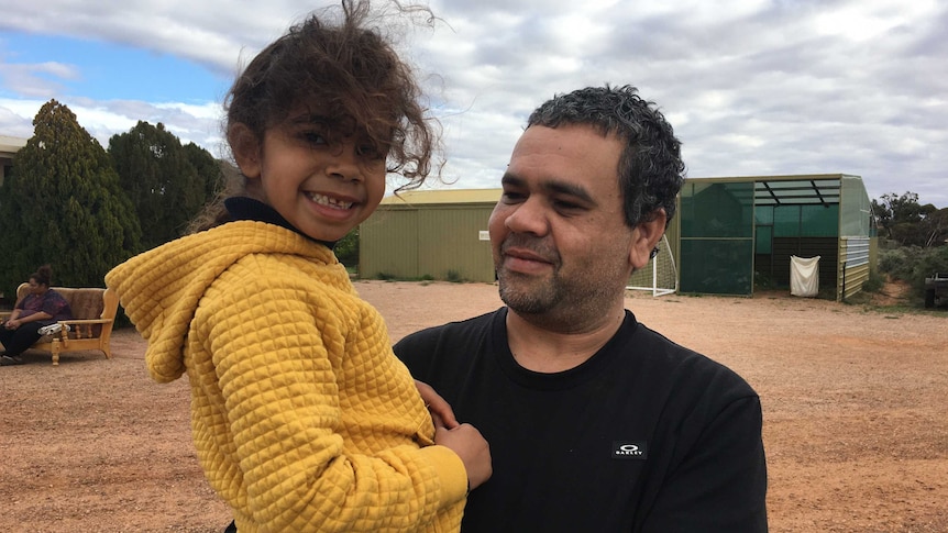 A young girl wearing a yellow hoodie, in the arms of her father Michael Donovan, grins towards the camera.