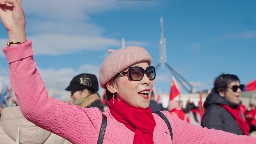 A smiling lady in a pink hat and top waves her hands as she stands in crowd in bright sun outside Federal Parliament.