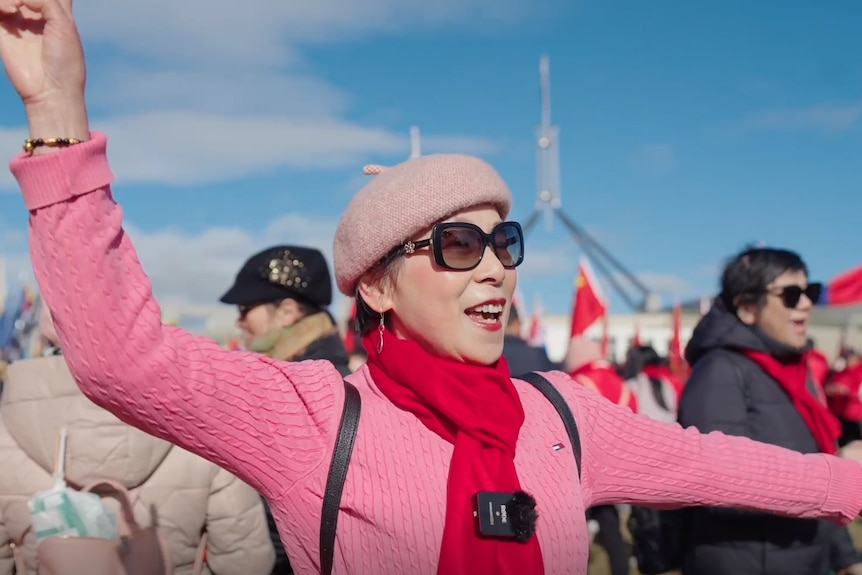 A smiling lady in a pink hat and top waves her hands as she stands in crowd in bright sun outside Federal Parliament.