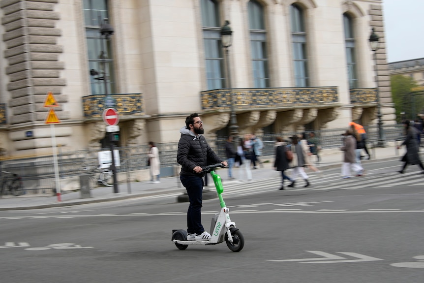 A bearded man without a helmet rides a green-and-white electric scooter along a road past an old stone building.