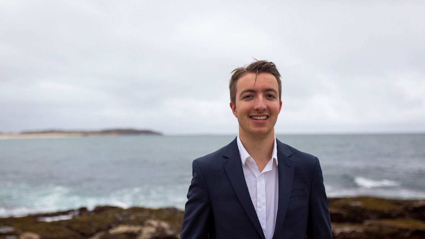 Declan Stelle, a Labor candidate, standing on some rocks overlooking the ocean. He is wearing a suit.