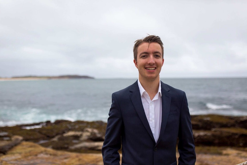 Declan Stelle, a Labor candidate, standing on some rocks overlooking the ocean. He is wearing a suit.