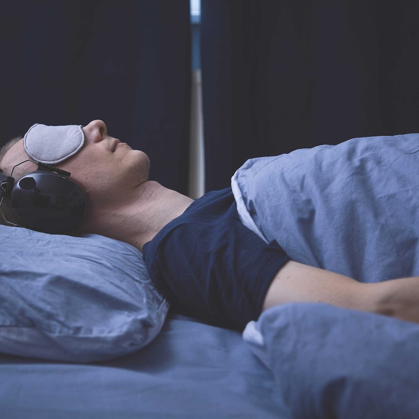 A man lying down on a bed in a dark room with an eyemask and headphones on